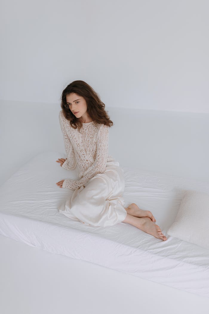 A woman in a cozy sweater sits thoughtfully on a bed in a minimalist room.